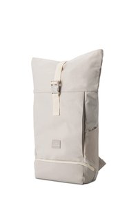 Sac à dos rectangulaire beige clair avec fermeture à boucle sur le dessus, écusson du logo à l'avant, poche latérale et compartiment à fermeture éclair en bas sur fond blanc.