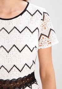 Vit och svart spetsblus med ett zigzagmönster, korta ärmar, rund halsringning och en svart spetsdetalj i midjan.
