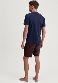 Navyblauwe fitted t-shirt en patroonshorts in donkerrood met een geometrisch ontwerp, gemaakt van lichtgewicht stof. Blootsvoets, gezien van achteren.