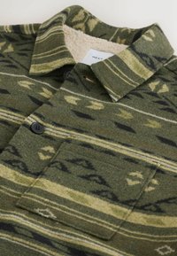 Green wool shirt featuring a textured design with geometric patterns in dark green, yellow, and black. Includes a collared neck and pocket.