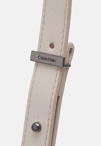 Biała skórzana bransoleta do zegarka z ozdobnym metalowym zapięciem w kolorze srebrnym, na którym wygrawerowano "Calvin Klein", oraz ozdobnym detalem w postaci guzika.