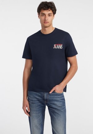 T-shirt en coton bleu marine à manches courtes, col rond, avec un logo graphique blanc et rouge sur la poitrine gauche. Porté avec un jean bleu clair.