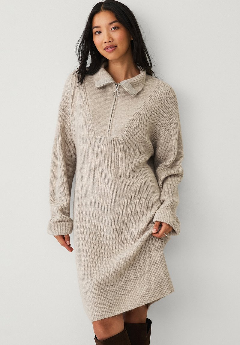 Beige gebreide jurk met een ritskraag, lange mouwen, geribbelde textuur, V-vormig paneel en een iets oversized pasvorm.