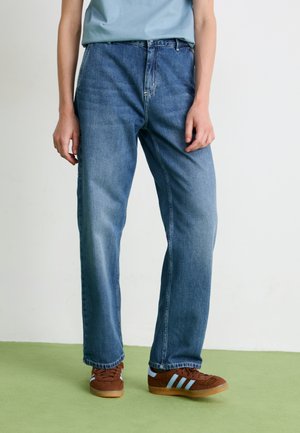 Person som har på seg avslappede blå jeans og brune joggesko med lyseblå striper, stående på et grønt gulv mot en hvit bakgrunn.