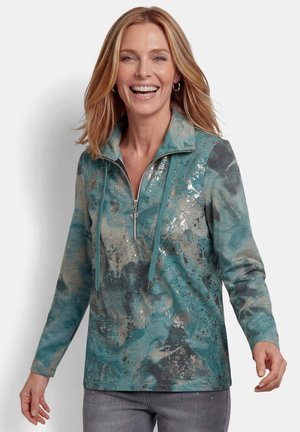 Lächelnde Frau, die einen türkis- und grauen Langarmpullover mit Reißverschluss und Kordelzug trägt, kombiniert mit grauen Hosen, vor einem schlichten Hintergrund stehend.