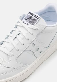 Zapatilla de cuero blanca con acentos texturizados, que presenta un cierre de cordones, punta redondeada y logotipo distintivo en la lengüeta.