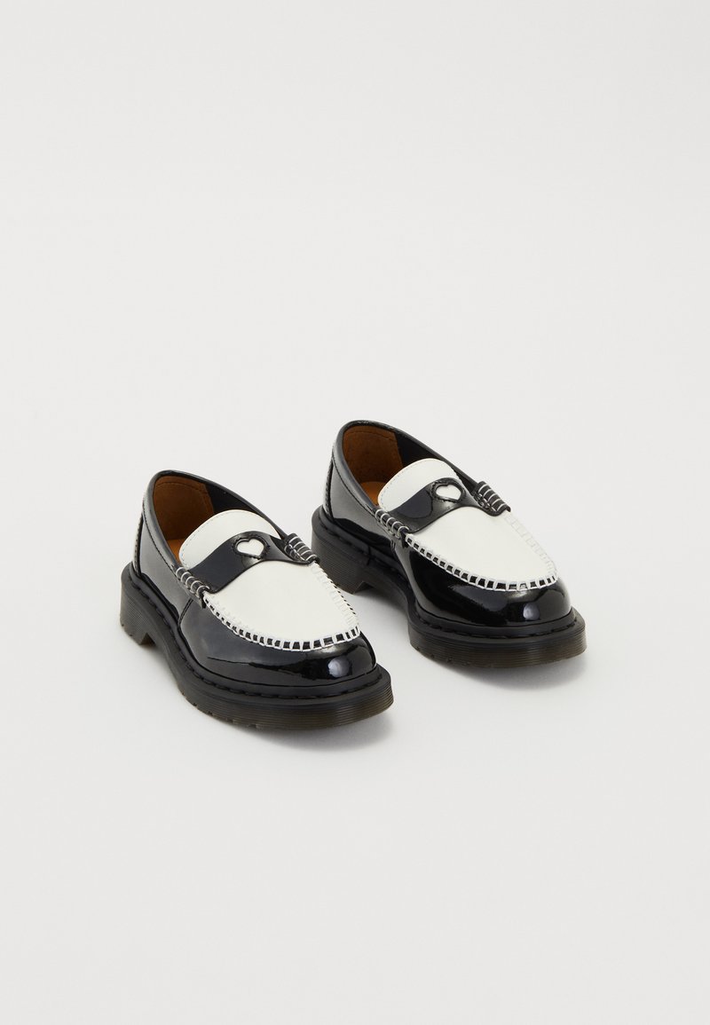 Melnbaltie ādas loaferi ar sirdī līdzīgu metāla detaļu augšpusē un biezām melnām zolēm, tie ir parādīti blakus uz baltā fona.