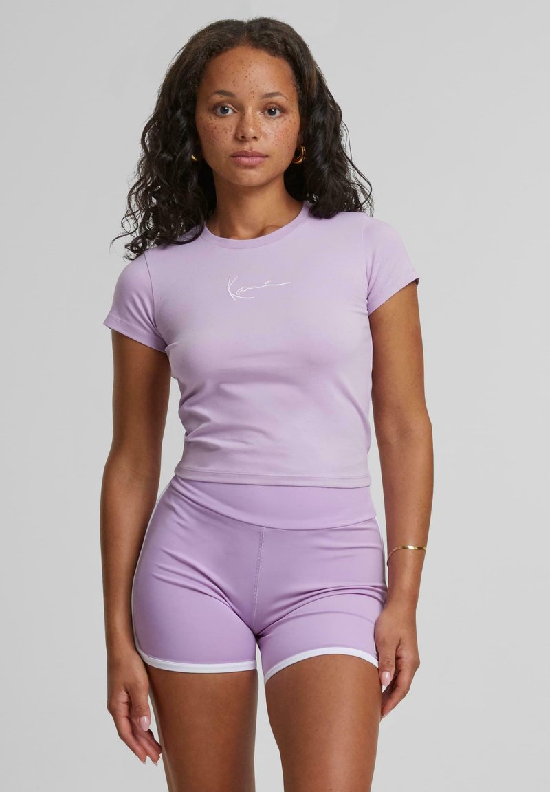 T-shirt viola aderente con maniche corte e un logo bianco, abbinato a pantaloni corti a vita alta dello stesso colore con rifiniture bianche. Tessuto dalla texture liscia.