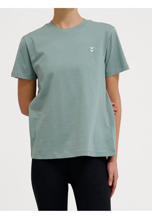 Person trägt ein hellgrünes T-Shirt mit kurzen Ärmeln und einem kleinen weißen Doppel-Pfeil-Logo auf der Brust und schwarze Hose.