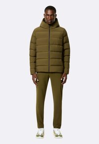 Giacca a vento verde oliva con trapuntatura verticale e cappuccio, abbinata a pantaloni coordinati e sneakers leggere con accenti verdi.