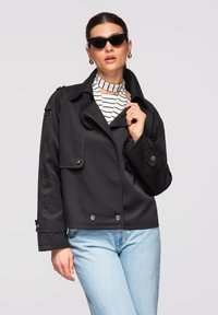 Femme portant une veste noire oversize, un haut à col montant rayé, un jean bleu clair, des lunettes de soleil œil-de-chat et des boucles d'oreilles créoles en or, posant devant un fond gris.