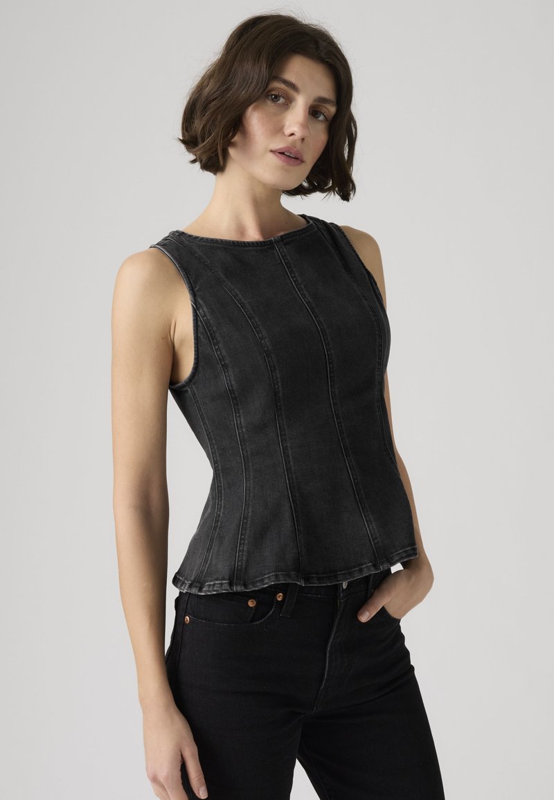 Woman wearing a fitted sleeveless black denim top paired with black jeans, standing against a plain light gray background.