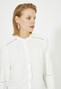 Witte button-up blouse met subtiele stikdetails langs de schouders en mouwen, voorzien van een ronde kraag en doorzichtige knopen.