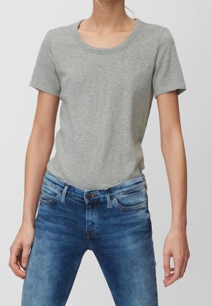 Person trägt ein eng anliegendes, hellgraues T-Shirt mit kurzen Ärmeln, das in eine mittelhohe blaue Jeans gesteckt ist, vor einem schlichten Hintergrund.