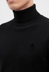 Homme portant un pull à col roulé côtelé noir, montrant le cou et le haut de la poitrine sur un fond uni.