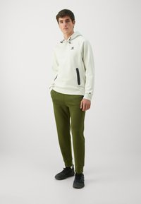 Hellgrauer Kapuzenpullover mit Logo und schwarzen Akzenten, kombiniert mit dunkelgrünen, taillierten Hosen und schwarzen Sneakern. Glattes Material, lässiges Design.