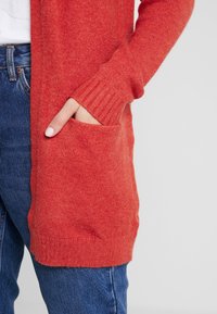 Röd stickad cardigan med ribbade ärmar och öppen front, med en sidoficka. Bärs över blå denimjeans.