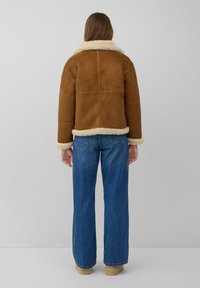 Bruine suède jas met een witte bontkraag en manchetten, met een boxy pasvorm, naden aan de achterkant, gedragen met blauwe brede jeans en beige laarzen.