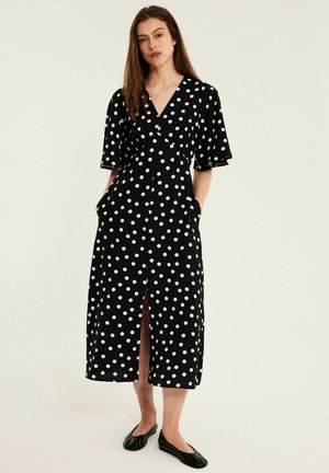 Kobieta stoi w czarnej sukience midi w białe kropki, z krótkimi rękawami, z rozporkiem z przodu i rękami w kieszeniach, nosząca czarne baleriny.