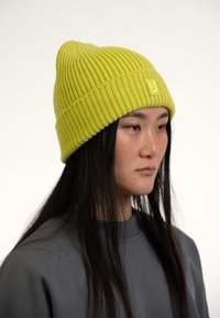 Gorro de punto verde lima con textura acanalada y un parche de logo en el centro. Llevado con una sudadera gris, que presenta una silueta relajada.
