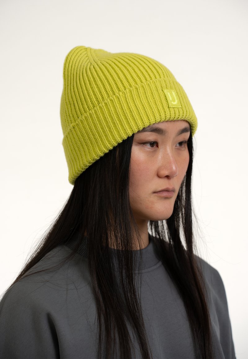 Gorro de punto verde lima con textura acanalada y un parche de logo en el centro. Llevado con una sudadera gris, que presenta una silueta relajada.