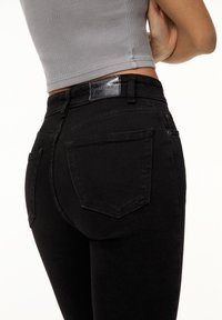 Jean skinny noir taille haute, avec une étiquette de marque brillante, deux poches arrière, et une texture lisse.