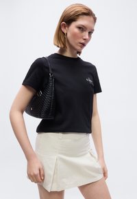 Νέα γυναίκα που φοράει μαύρο t-shirt Calvin Klein, μπεζ πλισέ μίνι φούστα, κρατώντας μαύρη υφασμάτινη τσάντα ώμου, με απλό φωτεινό φόντο.