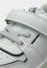 Λευκό αθλητικό παπούτσι με λείο συνθετικό υλικό, γκρι κορδόνια, λουράκι με βέλκρο και διακριτικές μαύρες λεπτομέρειες κατά μήκος της ραφής και της μύτης.