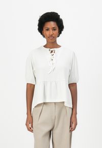 Weißes Peplum-Top mit rundem Ausschnitt, Schnürdetail an der Vorderseite und gepufften Ärmeln. Glatte Textur kombiniert mit beigen Hosen.