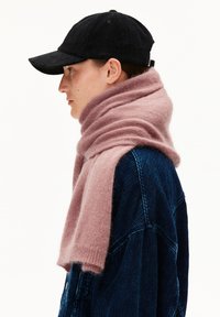 Persona di profilo che indossa un cappellino nero, una giacca di velluto a coste blu oversized e una sciarpa spessa di colore rosa chiaro avvolta attorno al collo.