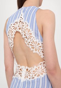 Femme portant une robe sans manches rayée bleu et blanc avec des découpes en dentelle florale blanche laissant apparaître le haut et le bas du dos.