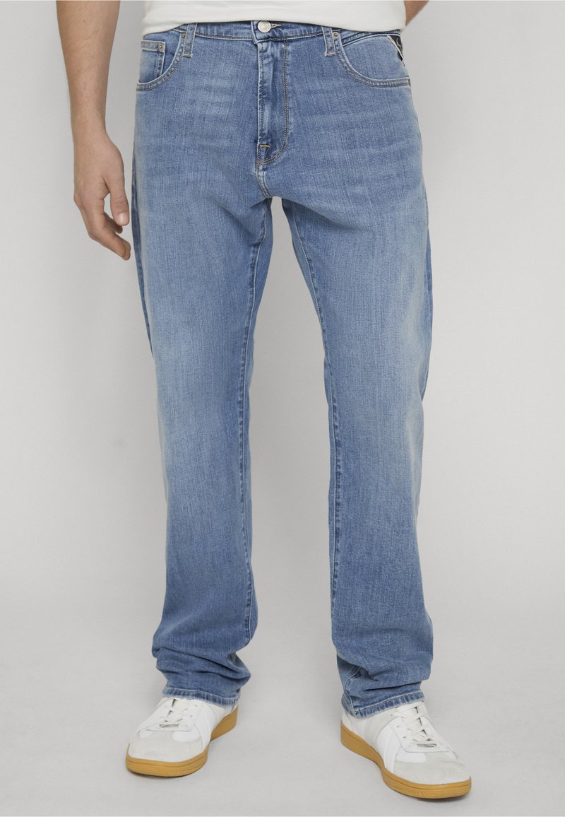 Homme portant un jean droit bleu clair et des baskets blanches, debout devant un fond uni gris clair.