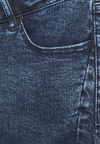 Tissu en denim bleu foncé avec une poche avant et des coutures visibles. La texture est légèrement usée avec un éclat lisse.