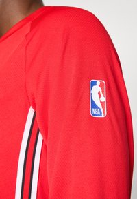 Manica di una maglia da sport rossa con strisce nere e bianche e toppa con il logo NBA sulla parte superiore del braccio.