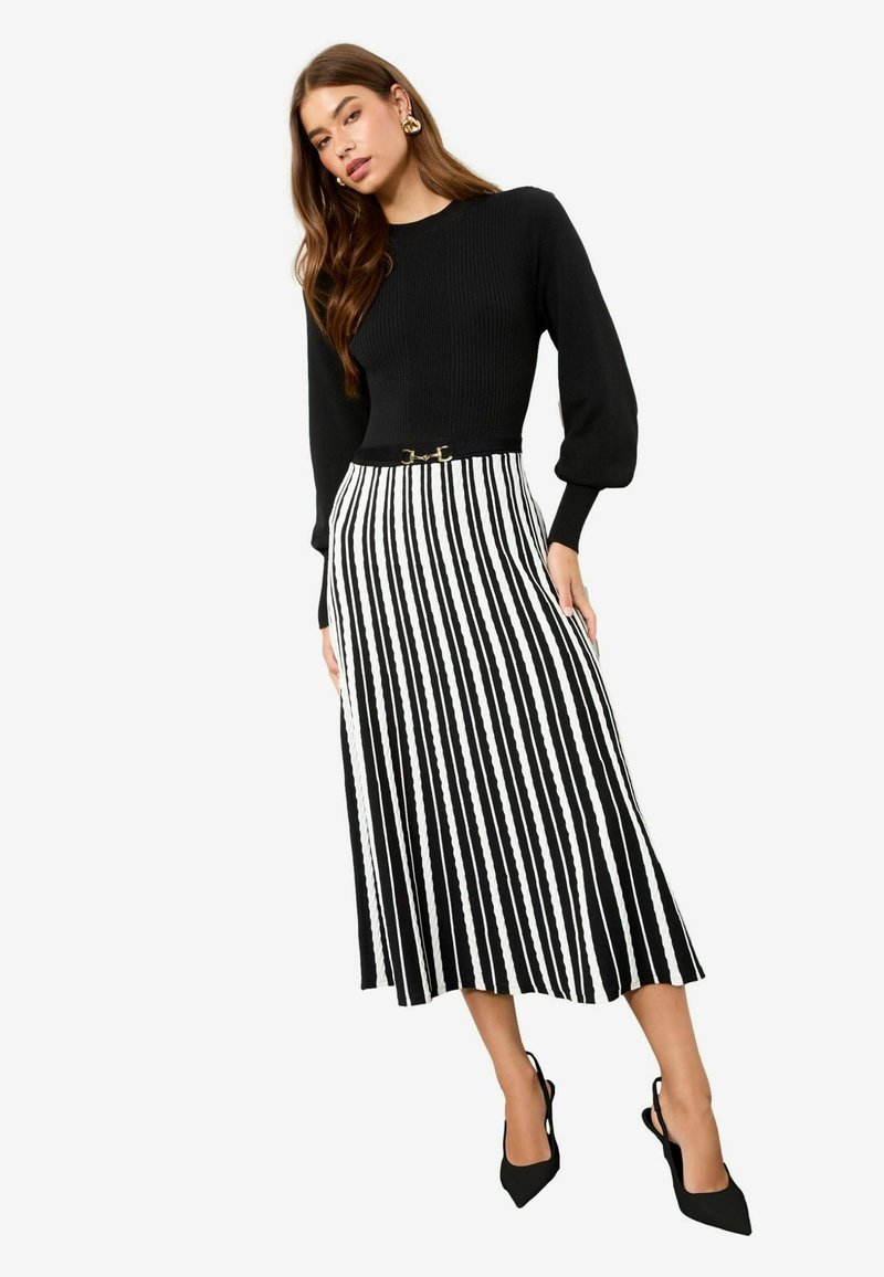 Melna ribota džemperis ar garām pūkām, kombinēts ar melnu un baltu svītru plisētu svārku. Svārkiem ir zelta akcentējošs josta.