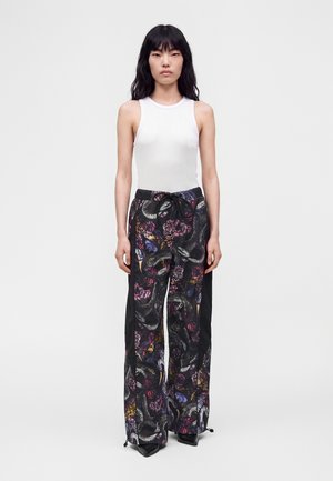 Femme asiatique portant un haut blanc sans manches et un pantalon noir à jambes larges avec des motifs colorés de serpent et de fleurs, debout devant un fond blanc.