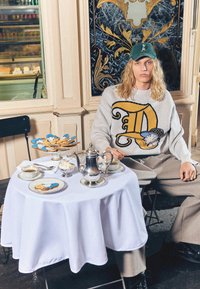 Pulover v svetlo sivi barvi z velikim rumenim motivom "D", sedeč za okroglo mizo z belo prtom, srebrno čajnik in okrašeni piškoti.