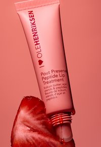 Rosa Tube von Ole Henriksen Pout Preserve Peptide Lip Treatment, mit rotem Text. Neben einer frischen Erdbeere platziert, mit Flüssigkeitstropfen.