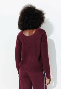 Pull en tricot bordeaux avec un large col rond et une texture côtelée, doté de manches longues et d'une coupe décontractée. Porté avec un pantalon assorti.