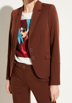 Femme portant un costume marron sur mesure avec un blazer à double boutonnage et un pantalon assorti, associé à un T-shirt blanc avec un motif graphique coloré représentant un visage.