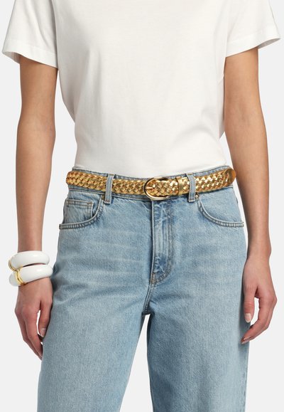 Person wearing light blue jeans with a woven gold belt, white short-sleeve shirt, and white and gold chunky bracelets on left wrist.