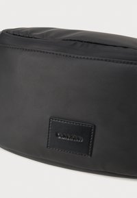 Fekete Calvin Klein táska sima, matt anyagból. Téglalap alakú, varrott bőrből készült logóval.