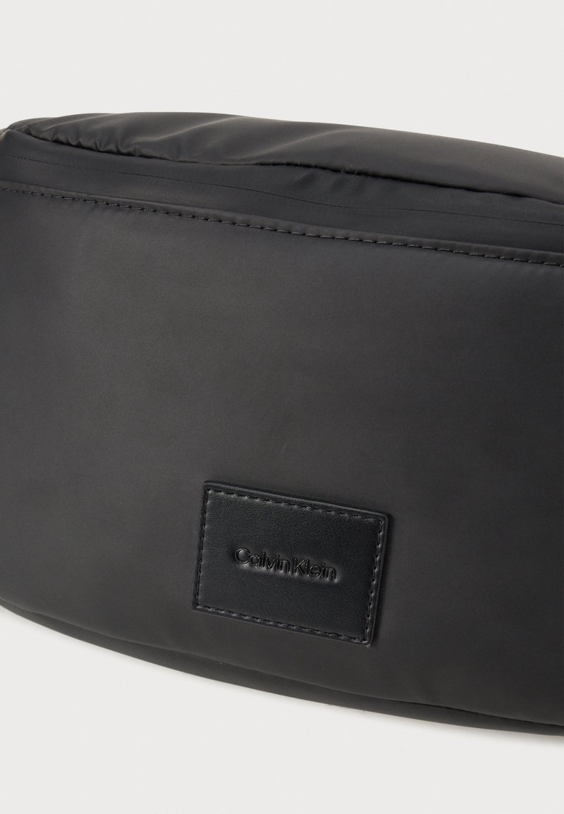 Sac noir Calvin Klein en tissu lisse et mat. Présente une forme rectangulaire avec un patch logo en cuir cousu.