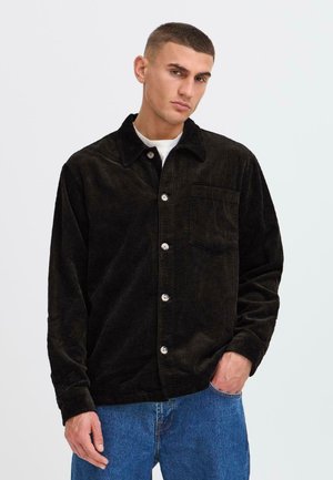 Chaqueta de camisa de pana negra con cuello tipo solapa, botones frontales y un bolsillo en el pecho, combinada con jeans azules. Se aprecia el detalle de la textura del tejido.
