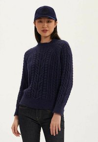 Pull en maille bleu marine avec un motif de tresses texturé, col et ourlet côtelés. Associé à une casquette bleue marine assortie, présenté avec un jean sombre.