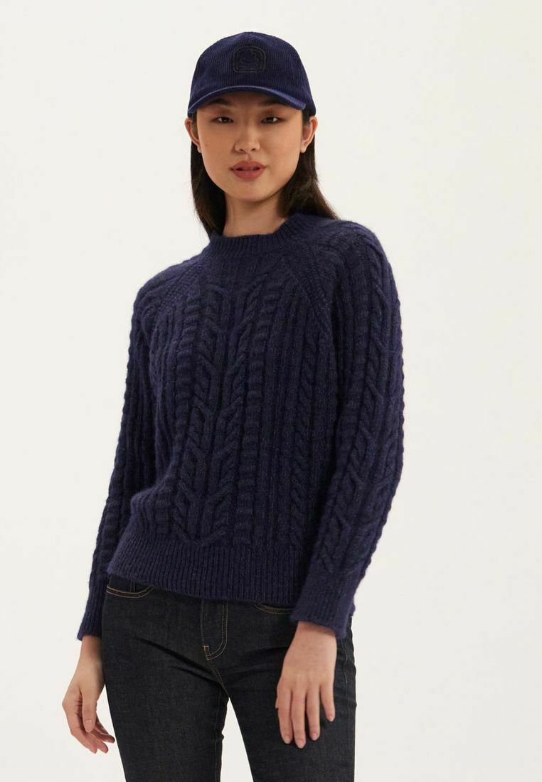 Pull en maille bleu marine avec un motif de tresses texturé, col et ourlet côtelés. Associé à une casquette bleue marine assortie, présenté avec un jean sombre.