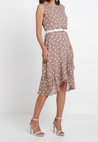 Braunes Polka-Dot-Kleid mit einem Rüschensaum, ausgestattet mit einem weißen Gürtel und dünnen Trägern, kombiniert mit weißen Knöchelriemchen-Absätzen.