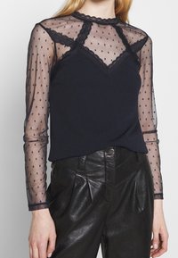 Långärmad topp med en marinblå enfärgad bas, en transparent prickig meshöverdrag och spetsdetaljer vid halsringningen och axlarna.