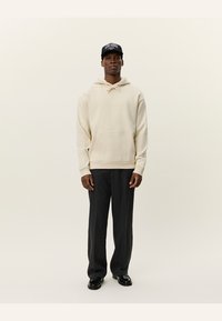 Crème hoodie met een kangoeroezak, gecombineerd met donkergrijze broeken. Het model draagt een zwarte pet met blauwe letters. Glad textuur van de stof.