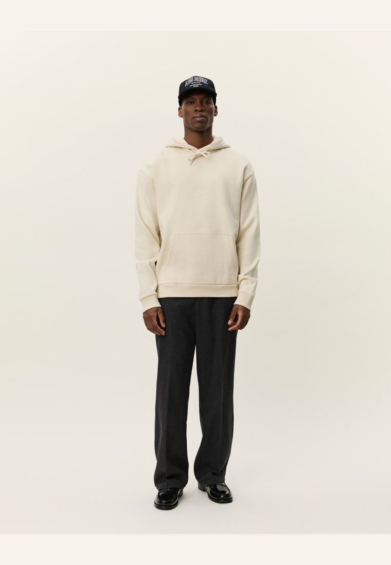 Crème hoodie met een kangoeroezak, gecombineerd met donkergrijze broeken. Het model draagt een zwarte pet met blauwe letters. Glad textuur van de stof.